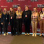 Group of eleven attendees standing shoulder to shoulder in front of a red conference backdrop with event branding, wearing business and business casual attire and conference name badges at an indoor venue. 