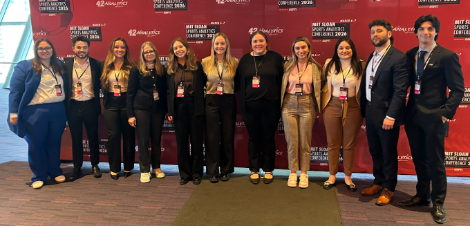 Group of eleven attendees standing shoulder to shoulder in front of a red conference backdrop with event branding, wearing business and business casual attire and conference name badges at an indoor venue. 