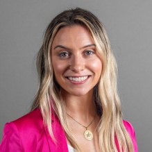 Portrait of Mary Starr wearing a hot pink jacket, white top, gold necklace, with a grey background. 