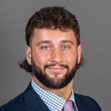 Portrait of Jon Stevens wearing dark grey jacket, blue checkered shirt and lavender tie, against a grey backdrop. 