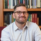 Robert Stark sits in a cushioned chair in front of bookshelves in a library, wearing glasses and a long sleeve shirt, with books visible on shelves behind him. 