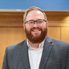 George Brown stands in a hallway of an academic building, wearing a suit jacket and glasses. 