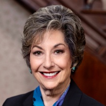 A professional portrait of an Ilene Fleischmann wearing a dark blazer and a patterned blue scarf, with a decorative brooch on the jacket. The individual is photographed indoors against a softly blurred background with warm tones. 
