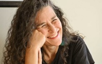 Close-up of a person with long curly hair resting their head on one hand, seated near a window with warm, soft lighting on the face and a neutral background. 