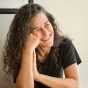 Close-up of a person with long curly hair resting their head on one hand, seated near a window with warm, soft lighting on the face and a neutral background. 