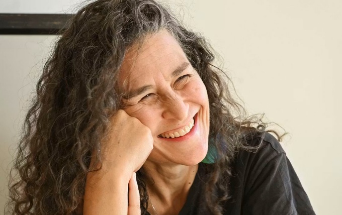 Close-up of a person with long curly hair resting their head on one hand, seated near a window with warm, soft lighting on the face and a neutral background. 