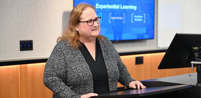 Person standing at a lectern in a classroom or conference room, speaking while a presentation slide is displayed on a screen behind them. The slide includes the title “Experiential Learning” with additional text beneath it. 