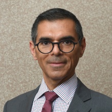 Portrait of a person wearing glasses and a suit with a red tie against a neutral background. 