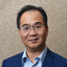 Portrait of a person wearing glasses and a blue blazer with a checkered shirt against a neutral background. 