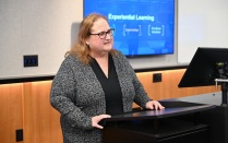 Person standing at a lectern in a classroom or conference room, speaking while a presentation slide is displayed on a screen behind them. The slide includes the title &ldquo;Experiential Learning&rdquo; with additional text beneath it. 