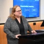 Person standing at a lectern in a classroom or conference room, speaking while a presentation slide is displayed on a screen behind them. The slide includes the title &ldquo;Experiential Learning&rdquo; with additional text beneath it. 