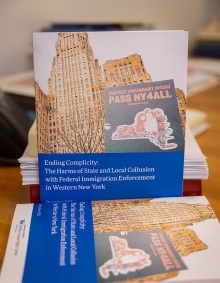 Stack of printed reports on a desk, with the top document showing a photo of a tall building and the title “Ending Complicity: The Harms of State and Local Collusion with Federal Immigration Enforcement in Western New York.” Books and papers are visible in the background. 