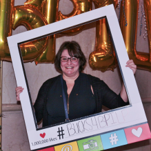 Dawn standing indoors holding a large decorative photo frame prop in front of gold balloon letters. The display includes greenery on either side and printed text on the frame that reads “# BUCKSL4BPILP” with social media style icons along the bottom. 