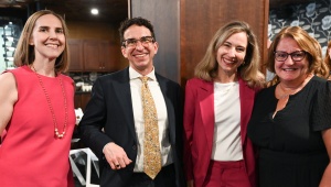 Four people standing together indoors at a reception or social event, dressed in business attire. The setting features wood paneling, wall decor, and a table with refreshments, with other attendees visible in the background. 