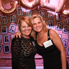 Two people stand together indoors with arms around each other at a formal event. Behind them is a brick wall decorated with gold balloon letters spelling “GOLD,” along with small framed photos and clusters of gold balloons. 