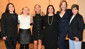 Six people stand side by side indoors at a formal event, with arms around each other. They are dressed in business and semi formal attire and pose on a patterned carpet in a brightly lit room with wall sconces. 