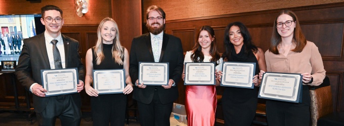 Seven people stand side by side indoors holding framed certificates, posed for a group photo. The individuals are dressed in formal or business attire and stand against a wood paneled wall in a dimly lit event space, suggesting an awards or recognition ceremony.