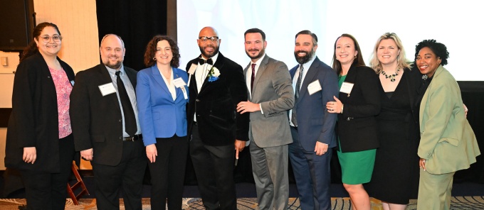 Nine attendees wearing formal or business attire stand together for a group photo at an indoor awards event, with a stage and large screen behind them. 