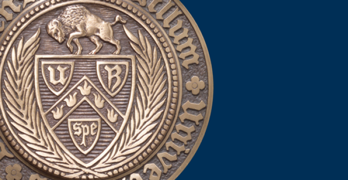 Partial close up of a bronze University at Buffalo seal featuring a buffalo and shield design, shown against a solid blue background. 