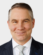 A person wearing a dark pinstripe suit, white dress shirt, and light patterned tie, posed against a plain light gray background. 