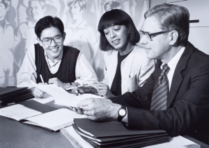 Black and white photo of two students with Prof. Schlegel looking over papers on a table.