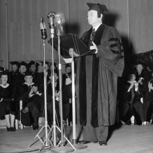 An old black and white image of Justice Robert H. Jackson standing on a stage with several microphones, reading a lecture to a group of peope. All are wearing graduation regalia. 