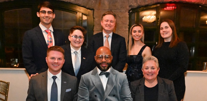 A group of nine people pose indoors in two rows at a formal event. Three individuals in the front row are seated and holding award plaques, while six others stand behind them. The group is dressed in formal attire, including suits, ties, and evening wear. The setting features stone archways, large windows, and warm ambient lighting. 