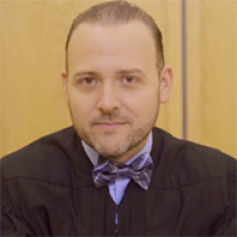 A person wearing a judicial robe and a patterned bow tie sits indoors in front of a light‑colored wooden wall. 