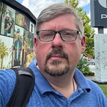 A person wearing a blue collared shirt and carrying a backpack stands outdoors near a wall with large photo murals and signage, with trees visible in the background. 