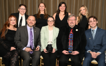 Zoom image: A group of nine individuals posed for a formal group photo. Three people are seated in the front row holding awards, and six are standing behind them. They are positioned in front of a floor‑length curtain, with a table and decorative candles to the right.