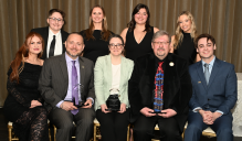 Zoom image: A group of nine individuals posed for a formal group photo. Three people are seated in the front row holding awards, and six are standing behind them. They are positioned in front of a floor‑length curtain, with a table and decorative candles to the right.