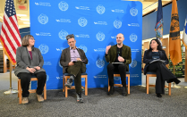 Zoom image: Four people seated in front of a blue University at Buffalo School of Law backdrop, participating in a panel discussion with U.S. and New York State flags displayed on each side.