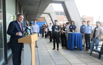 Zoom image: Person speaking at a podium during a networking event while attendees listen on an outdoor terrace.