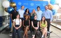 Zoom image: Group photo of nine students posing in front of a University at Buffalo School of Law backdrop and blue, white, and gold balloons at an outdoor event.