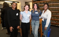 Zoom image: Four individuals stand side by side in a library‑style room with shelves of law books lining the walls. Each person is wearing clothing suitable for an academic or professional setting and has a name badge attached. Wooden tables and chairs are visible behind them, and the space is evenly lit indoors.