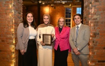 Zoom image: Four people standing indoors at an awards banquet, smiling and posing for a group photo. One person holds a framed award plaque, and all are dressed in formal or business attire. Exposed brick walls and event lighting are visible in the background.