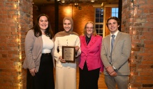 Zoom image: Four people standing indoors at an awards banquet, smiling and posing for a group photo. One person holds a framed award plaque, and all are dressed in formal or business attire. Exposed brick walls and event lighting are visible in the background.