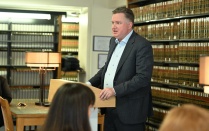 Zoom image: An individual stands behind a wooden lectern in a library‑style room lined with shelves of law books. Several seated people are visible in the foreground, facing the lectern. A table lamp and wooden furniture appear nearby, and the space is evenly lit with indoor lighting.
