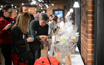 Zoom image: People gather around a long display table filled with gift bags and wrapped raffle items in a brick-walled event space.