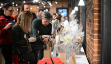 Zoom image: People gather around a long display table filled with gift bags and wrapped raffle items in a brick-walled event space.