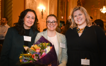 Zoom image: Three individuals standing together in an ornate event space with warm lighting, tall decorative columns, and chandeliers overhead. One person in the center is holding a large bouquet of colorful flowers wrapped in dark purple paper. Numerous attendees are visible in the background.