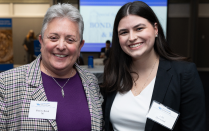 Zoom image: Two people standing together at an indoor event, one wearing a plaid blazer over a purple top and the other wearing a dark blazer over a white blouse. Both have name tags, and a presentation screen is visible in the background.