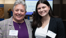 Zoom image: Two people standing together at an indoor event, one wearing a plaid blazer over a purple top and the other wearing a dark blazer over a white blouse. Both have name tags, and a presentation screen is visible in the background.