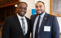 Zoom image: Two men in suits standing indoors at an alumni event, smiling and posing for a photo. One wears a name badge on his jacket, and bookshelves and framed artwork are visible in the background.