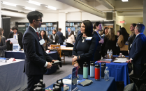 Zoom image: A job candidate speaks with an employer representative at a career fair inside a library. Tables covered with informational materials, water bottles, and branded items are arranged throughout the space. Other attendees and employers are visible having conversations in the background, with bookshelves lining the walls.