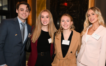 Zoom image: Four individuals standing together in a lobby‑style area with marble walls and framed artwork. They are dressed in professional attire, including suits and coordinated jackets. A doorway and additional event attendees are visible behind them.