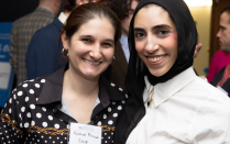 Zoom image: Two people standing close together and posing for a photo at an indoor event. One is wearing a black and white patterned shirt with a name tag, and the other is wearing a white top with a black headscarf. Other attendees are visible in the background.