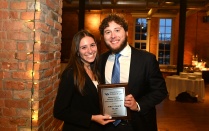 Zoom image: Two law students standing indoors at an awards banquet, smiling and holding a first place team award plaque. They are dressed in formal attire, with exposed brick walls and event tables visible in the background.