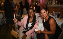 Zoom image: Indoor evening event with attendees seated at round tables covered in white tablecloths. Two people sit in the foreground holding lit candles, while other attendees stand and sit in the softly lit background. Candles, glassware, and place settings are visible on the tables, creating a formal or ceremonial atmosphere.