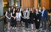 Zoom image: A large group of attendees stands closely together indoors, posing for a group photo at a formal networking event, with tall windows, curtains, and banquet seating visible in the background.